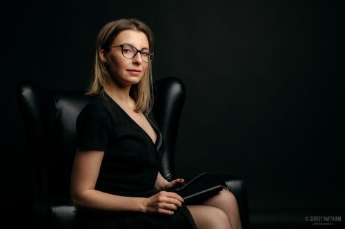 Деловой портрет женщины в очках и черном платье, сидящей в кожаном кресле для бизнес съемки