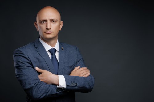 Лысый мужчина в синем деловом костюме и галстуке со скрещенными на груди руками на сером фоне